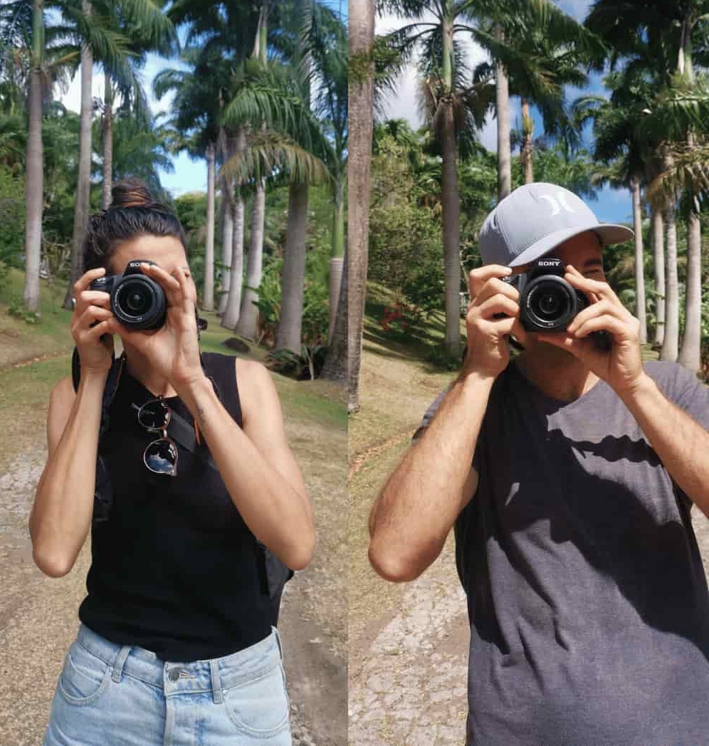 Katharina and Allan, the authors behind this travel blog called Happy Healthy Nomads, standing next to each other with palm trees in the background, holding DSLR cameras in front of their faces.